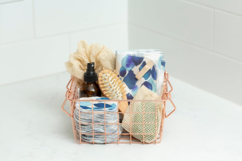 May include: A copper wire basket filled with bathroom essentials, including a reusable cotton round set, a natural hair brush, a mesh bag, a bottle of liquid, and a beige scrunchie.