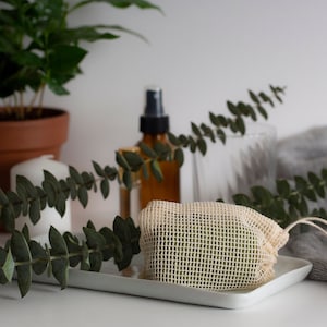 May include: A white mesh bag containing a green bar of soap, resting on a white tray. The bag is secured with a white string. In the background, there is a plant, a candle, and a glass bottle.