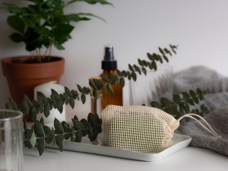 May include: A white mesh bag containing a green bar of soap, resting on a white tray. The bag is secured with a white string. In the background, there is a plant, a candle, and a glass bottle.