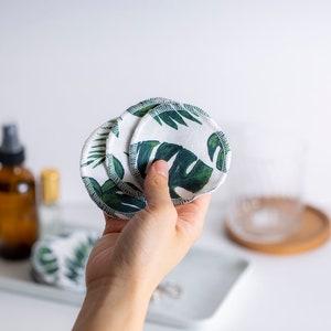 May include: A hand holding a stack of reusable cotton rounds with a green tropical leaf print. The rounds are white with green leaves and have a white stitched edge.
