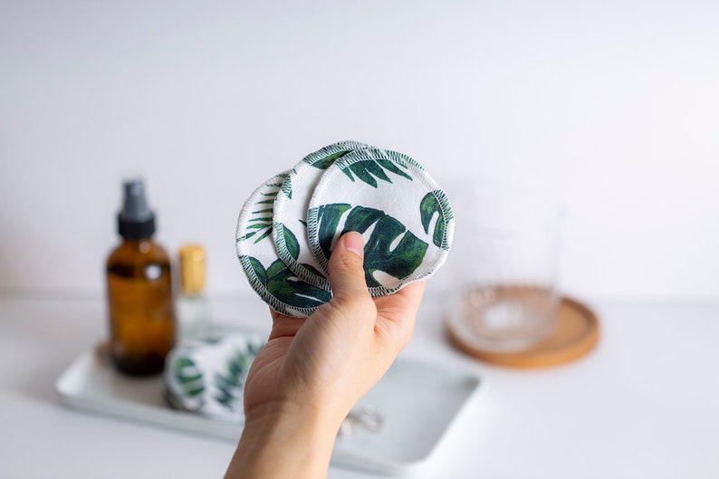 May include: A hand holding a stack of reusable cotton rounds with a green tropical leaf print. The rounds are white with green leaves and have a white stitched edge.
