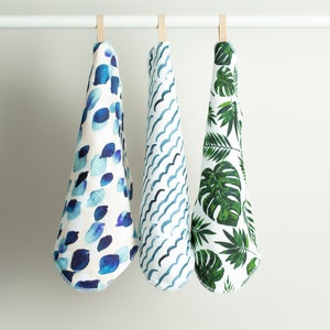 May include: Three kitchen towels hanging on a white rod. The towels are white with different blue and green patterns. The first towel has a blue watercolor pattern of circles. The second towel has a blue and white wavy pattern. The third towel has a green tropical leaf pattern.