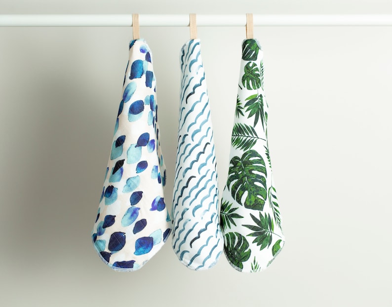 May include: Three kitchen towels hanging on a white rod. The towels are white with different blue and green patterns. The first towel has a blue watercolor pattern of circles. The second towel has a blue and white wavy pattern. The third towel has a green tropical leaf pattern.