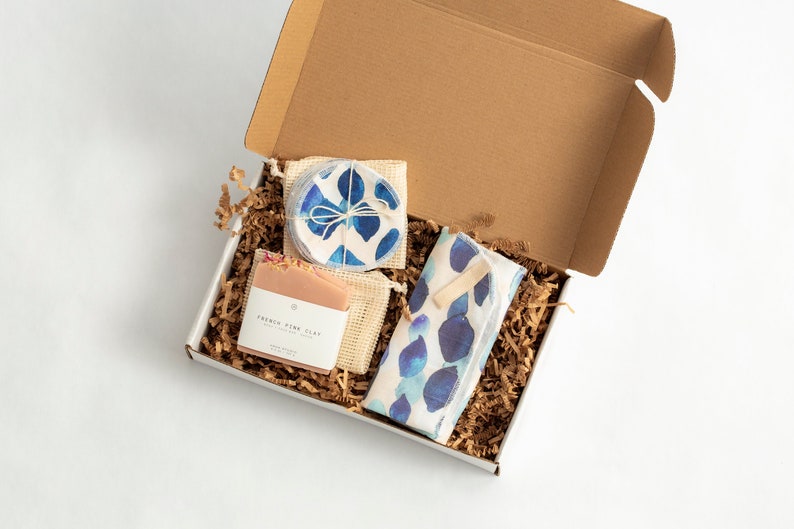 May include: A gift box filled with brown packing paper and containing a bar of soap labeled "French Pink Clay", a set of reusable makeup remover pads in a mesh bag, and a blue and white patterned cloth.
