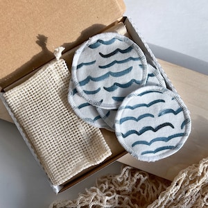 Könnte beinhalten: Wiederverwendbare Wattepads mit blauem Wellenmuster in einer Pappschachtel mit einem Netzbeutel. Die Pads sind weiß mit einem blauen Wellendesign. Die Schachtel und der Beutel sind beige.