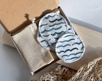 May include: A set of reusable cotton facial rounds with a blue wave pattern, presented in a cardboard box with a mesh bag. The rounds are white with a blue wave design. The box and bag are beige.