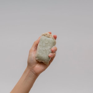 May include: A hand holding a beige and green soap sack. The soap sack is made of a mesh material and is filled with a white soap.