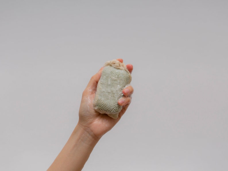 May include: A hand holding a beige and green soap sack. The soap sack is made of a mesh material and is filled with a white soap.