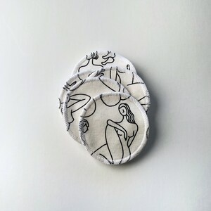 May include: A stack of five white reusable makeup remover pads with a black line drawing of a woman on each pad.