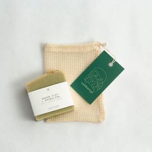 May include: A green bar of soap with a white label that reads "Green Clay + Spirulina Body + Face Bar Soap" is sitting on a beige mesh bag. A green tag with the word "wellbeing" and a leaf design is attached to the bag.