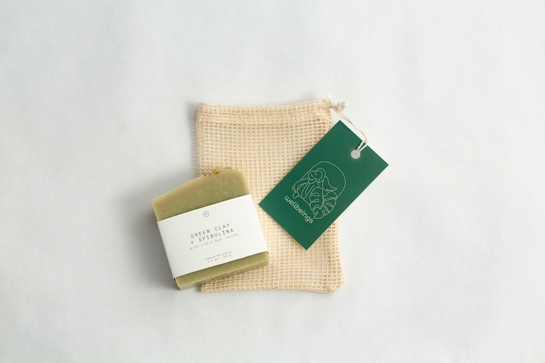 May include: A green bar of soap with a white label that reads "Green Clay + Spirulina Body + Face Bar Soap" is sitting on a beige mesh bag. A green tag with the word "wellbeing" and a leaf design is attached to the bag.