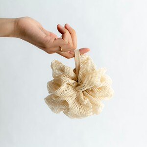 May include: A beige bath pouf with a beige elastic loop. The pouf is made of a soft, mesh material.