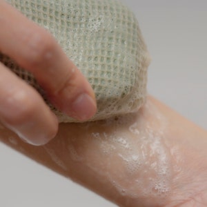 May include: A hand holding a green loofah sponge and rubbing it on a soapy arm.