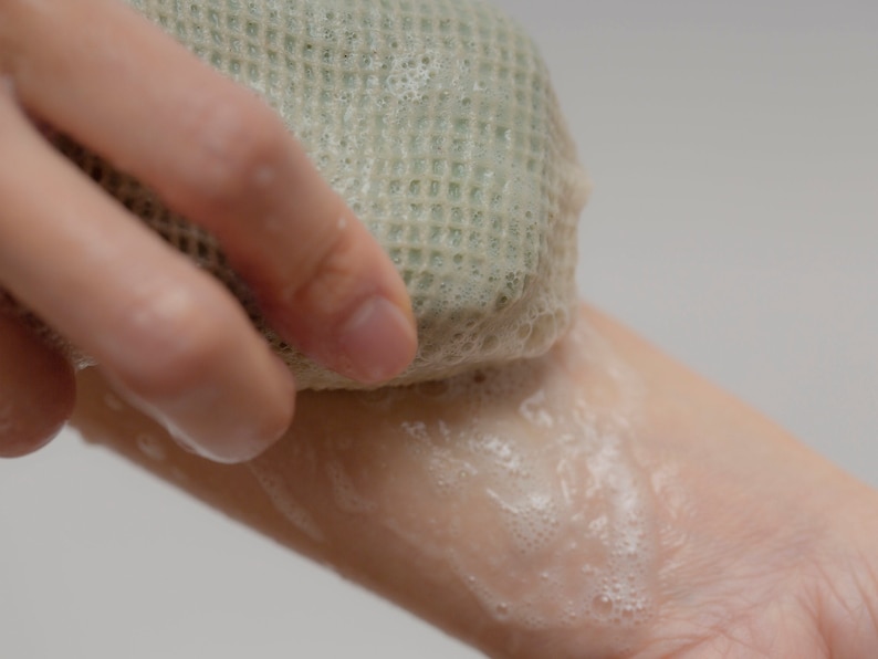 May include: A hand holding a green loofah sponge and rubbing it on a soapy arm.