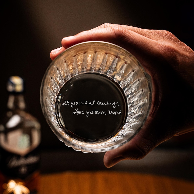 Puede incluir: Un vaso de vidrio transparente con una superficie texturizada. El vaso est&aacute; siendo sostenido por una mano y tiene las palabras "25 a&ntilde;os y contando... Te quiero m&aacute;s, Dixie" escritas en el interior.