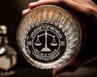 May include: A clear glass with a black and white engraved design. The design features a scale of justice, the words "University of Michigan", "20", "25", "Meet me at the bar!" and the name "Christian Brian".