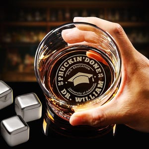 May include: A clear whiskey glass with the words "PHUCKIN' DONE" and "DR. WILLIAM" etched on the bottom. The glass is filled with amber liquid and held by a hand. Stainless steel ice cubes are in the foreground.
