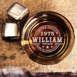 May include: A clear glass with a brown liquid inside. The glass has an engraved design that says "Limited Edition 1975 William Vintage Aged to Perfection".