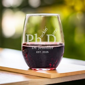 May include: A stemless wine glass with the text "Ph.D.  F*ckin' Done" and "Dr. Jennifer EST. 2025" etched on the side. The glass is filled with red wine.