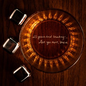 Puede incluir: Un vaso de whisky de color &aacute;mbar con un mensaje inscrito en el fondo: "25 a&ntilde;os y contando... Te quiero m&aacute;s, Dixie". El vaso est&aacute; rodeado de cuatro cubitos de hielo plateados.