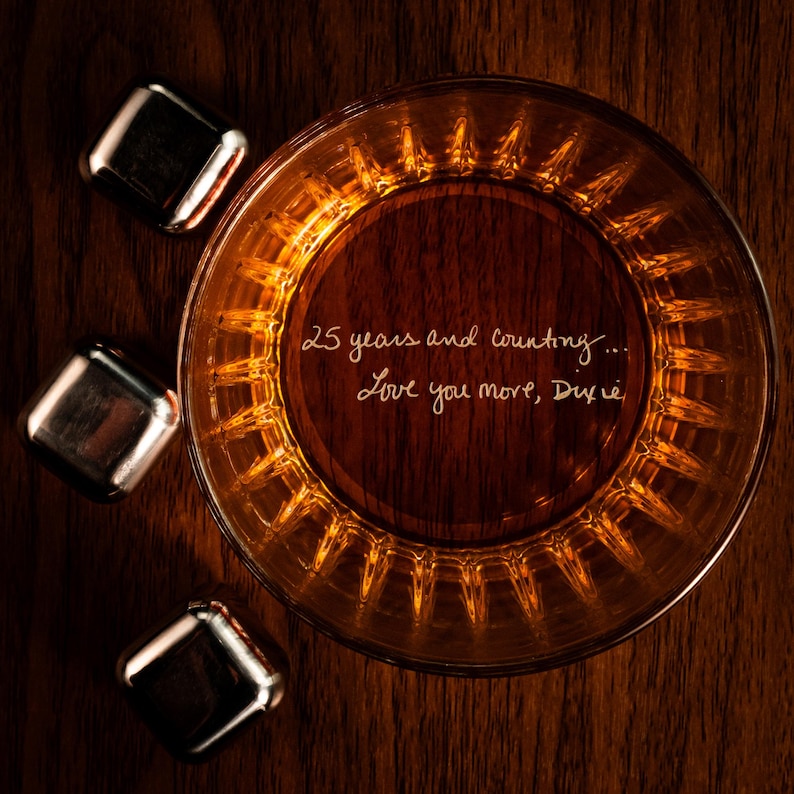 Puede incluir: Un vaso de whisky de color &aacute;mbar con un mensaje inscrito en el fondo: "25 a&ntilde;os y contando... Te quiero m&aacute;s, Dixie". El vaso est&aacute; rodeado de cuatro cubitos de hielo plateados.