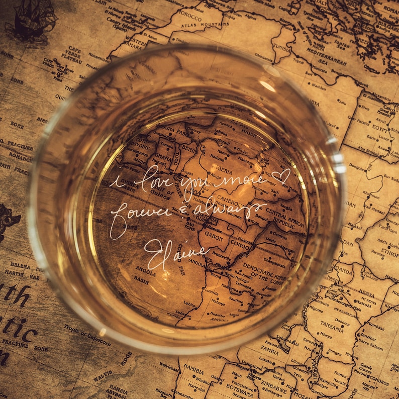 Puede incluir: Un primer plano de un mapa del mundo vintage con un vaso de l&iacute;quido encima. El mapa est&aacute; centrado en &Aacute;frica y el texto "I love you more forever & always Elaine" est&aacute; escrito en el vaso.
