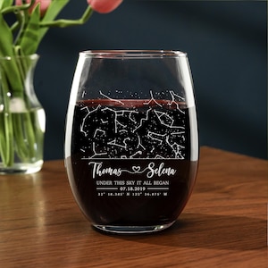 May include: A stemless wine glass filled with red liquid. The glass features a white constellation design and the names "Thomas & Selena" with the text "UNDER THIS SKY IT ALL BEGAN 07.18.2019 32° 18.385' N 122° 36.875' W". The glass sits on a wooden surface.
