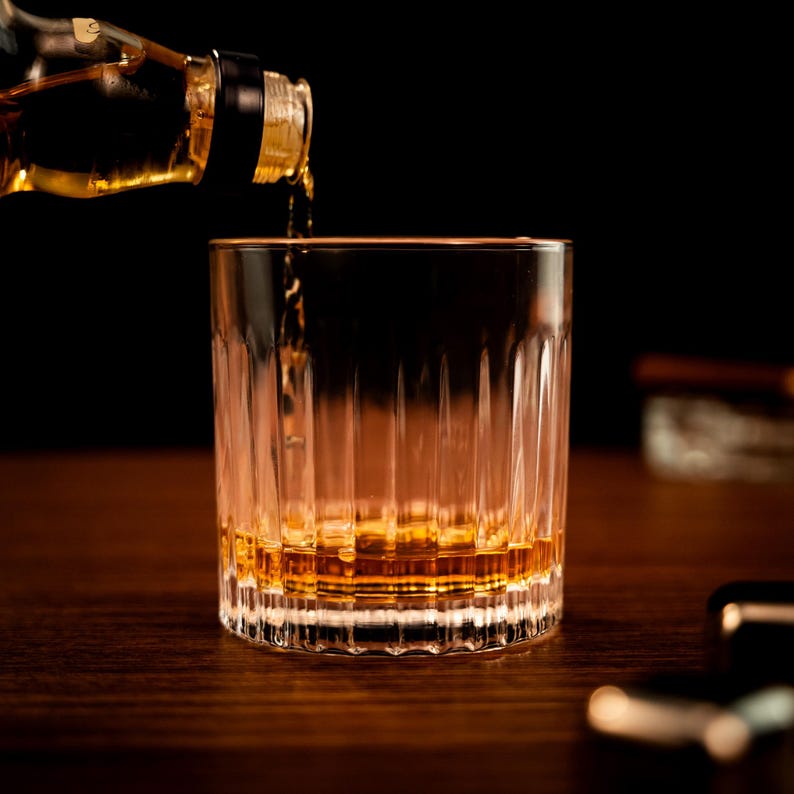 May include: A clear glass with a textured surface is being filled with amber-colored liquid from a bottle. The glass is sitting on a wooden surface.