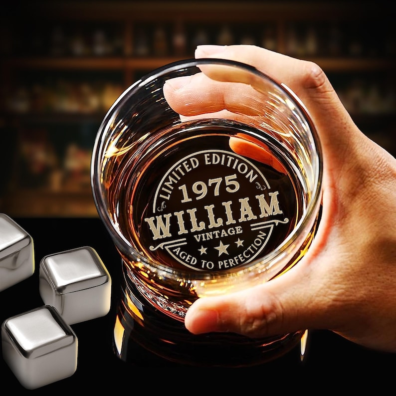 May include: A clear glass whiskey tumbler with an engraved design that reads "Limited Edition 1975 William Vintage Aged to Perfection". The glass is filled with amber liquid and is being held by a hand.