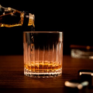 Puede incluir: Un vaso transparente con una superficie texturizada se est&aacute; llenando con l&iacute;quido de color &aacute;mbar de una botella. El vaso est&aacute; sobre una superficie de madera.