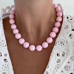 May include: A light pink beaded necklace with large, round beads. The necklace is worn against a white eyelet lace top. The beads are evenly spaced and appear to be made of a smooth material. The necklace is a statement piece.