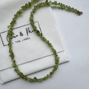 May include: A delicate necklace made of small, irregularly shaped, light green peridot stones. The necklace is displayed on a white cloth with the text "Clare & Me THE LABEL" in black. The clasp is gold-coloured.