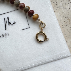 May include: A beaded bracelet featuring smooth, multi-colored stone beads in shades of red, brown, gray, and yellow, accented with small gold-toned spacers. The bracelet is finished with a gold-toned circular spring ring clasp and jump ring.