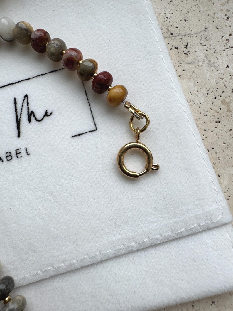 May include: A beaded bracelet featuring smooth, multi-colored stone beads in shades of red, brown, gray, and yellow, accented with small gold-toned spacers. The bracelet is finished with a gold-toned circular spring ring clasp and jump ring.