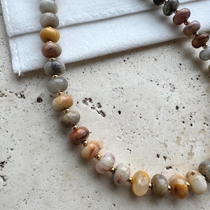 May include: A necklace featuring a series of smooth, multi-colored stone beads in shades of tan, brown, gray, and cream, interspersed with small, polished gold-tone metal spacer beads arranged in a curved line on a white fabric surface.