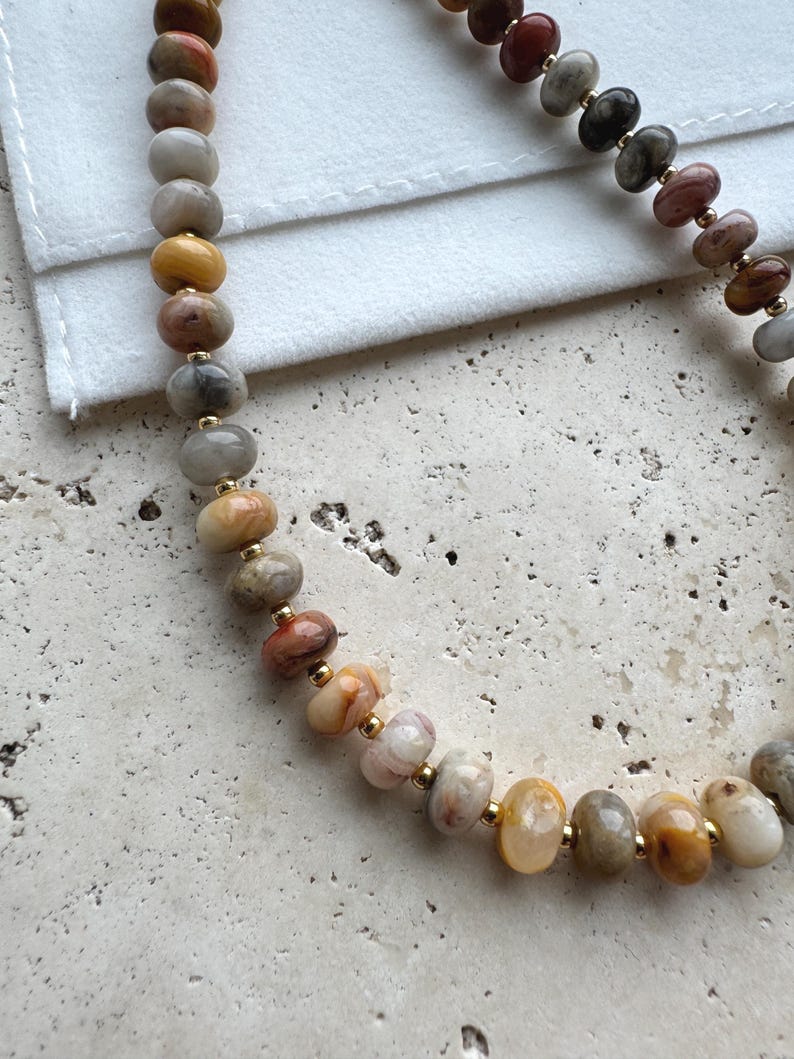 May include: A necklace featuring a series of smooth, multi-colored stone beads in shades of tan, brown, gray, and cream, interspersed with small, polished gold-tone metal spacer beads arranged in a curved line on a white fabric surface.