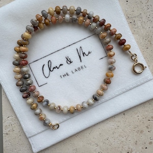 May include: A beaded necklace featuring small, round, multi-colored stone beads in shades of tan, gray, and reddish-brown, interspersed with tiny gold-toned metal spacers. The necklace has a gold-toned circular spring ring clasp at the end.