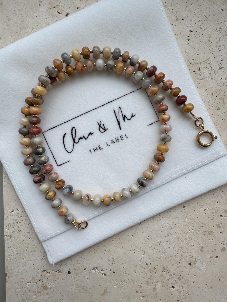 May include: A beaded necklace featuring small, round, multi-colored stone beads in shades of tan, gray, and reddish-brown, interspersed with tiny gold-toned metal spacers. The necklace has a gold-toned circular spring ring clasp at the end.