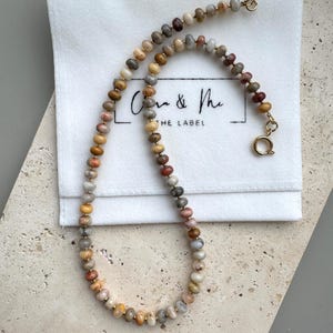 May include: A beaded necklace featuring small, round, multi-colored stone beads in shades of tan, gray, brown, and muted red, separated by tiny gold-toned spacers. The necklace includes a gold-toned circular spring ring clasp and a small jump ring.