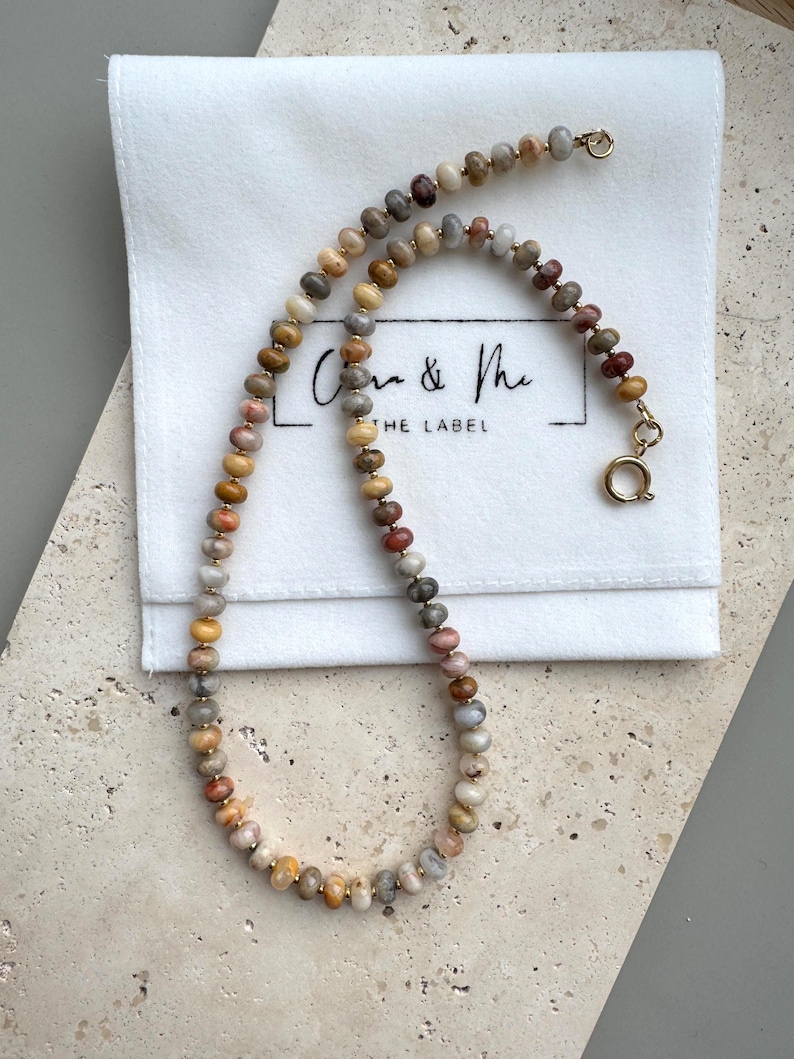May include: A beaded necklace featuring small, round, multi-colored stone beads in shades of tan, gray, brown, and muted red, separated by tiny gold-toned spacers. The necklace includes a gold-toned circular spring ring clasp and a small jump ring.