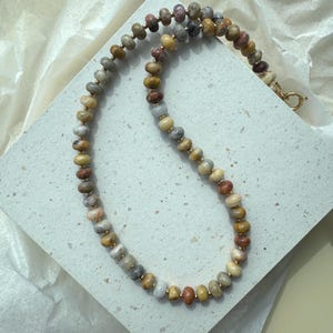 May include: A beaded necklace featuring round, multi-colored stone beads in shades of tan, brown, grey, and muted red. The beads are separated by small, metallic gold-toned spacers, and the necklace is secured with a gold-toned circular clasp.