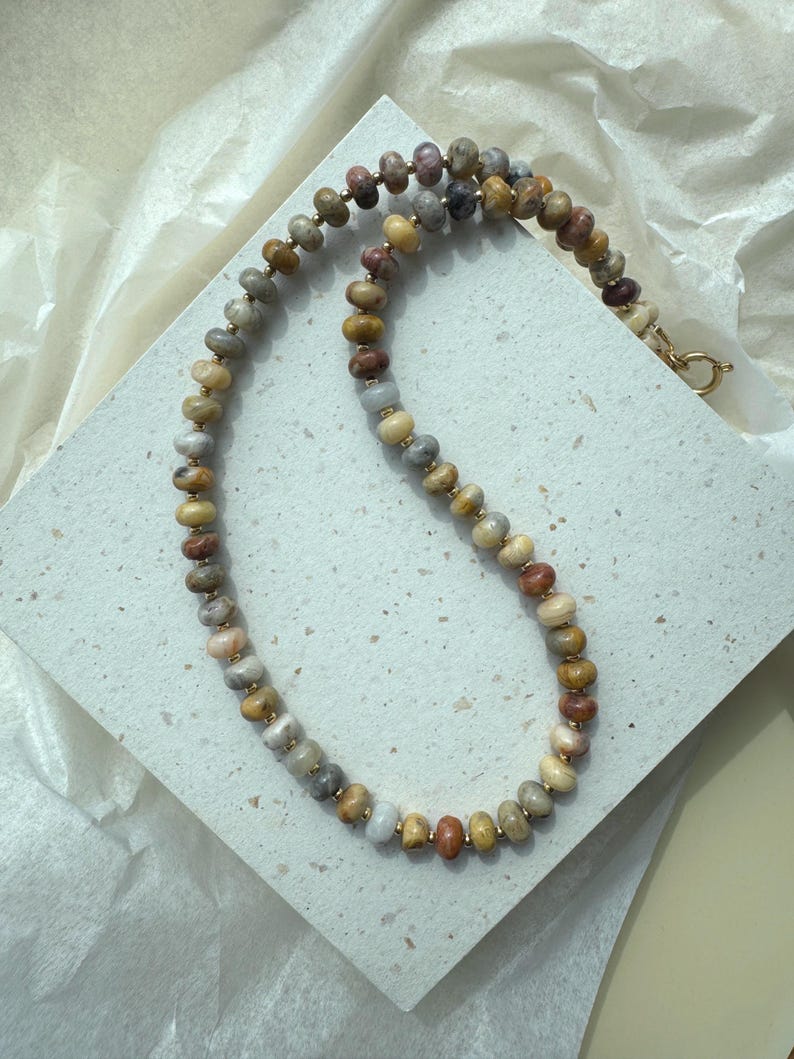 May include: A beaded necklace featuring round, multi-colored stone beads in shades of tan, brown, grey, and muted red. The beads are separated by small, metallic gold-toned spacers, and the necklace is secured with a gold-toned circular clasp.