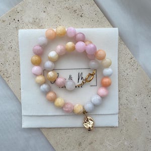 Könnte beinhalten: Eine mehrfarbige Perlenkette mit einem goldenen Anhänger. Die Perlen sind rosa, weiß und gelb. Die Kette liegt auf einem weißen Hintergrund.