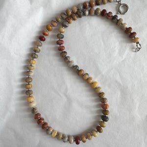 May include: A beaded necklace featuring small, round, multi-colored stone beads in shades of tan, brown, gray, and reddish-orange. The beads are separated by tiny silver-toned metal spacers and the necklace is secured with a silver-toned spring ring clasp.