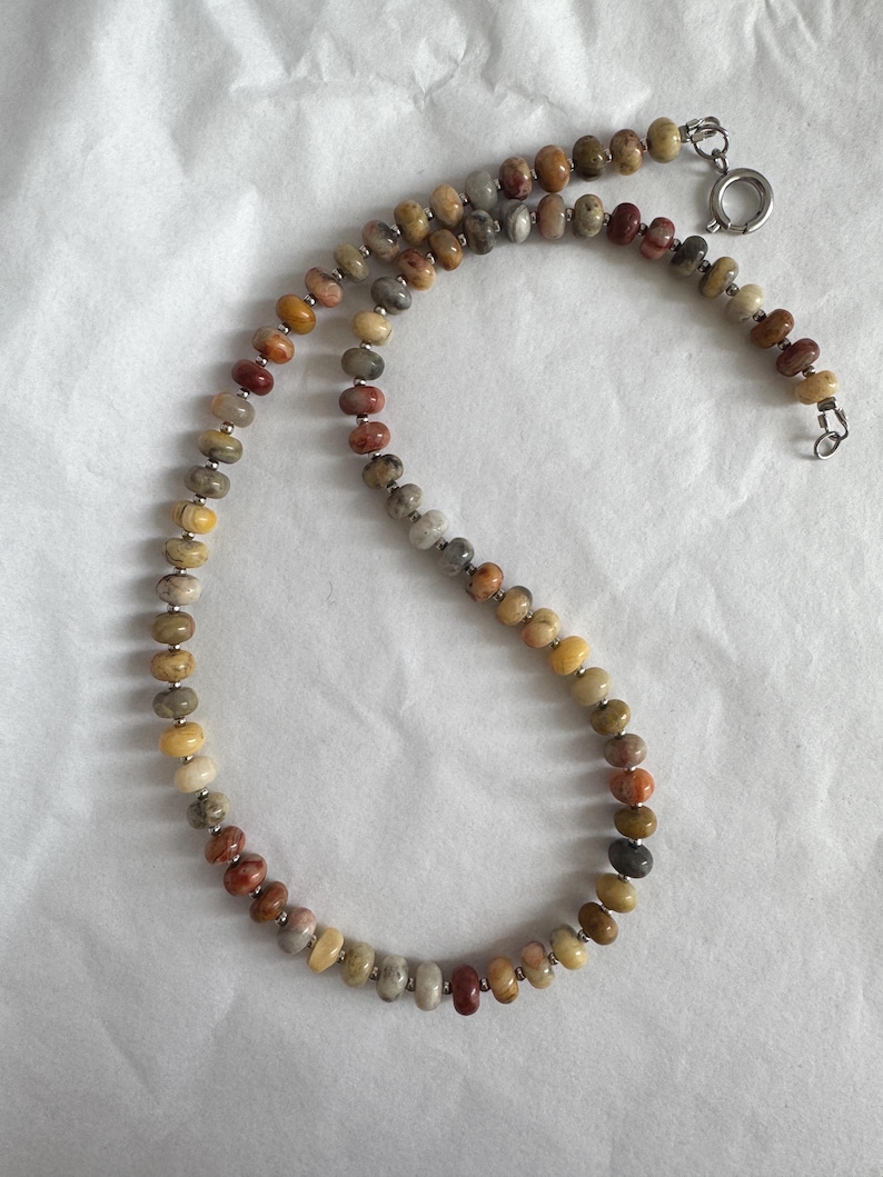 May include: A beaded necklace featuring small, round, multi-colored stone beads in shades of tan, brown, gray, and reddish-orange. The beads are separated by tiny silver-toned metal spacers and the necklace is secured with a silver-toned spring ring clasp.