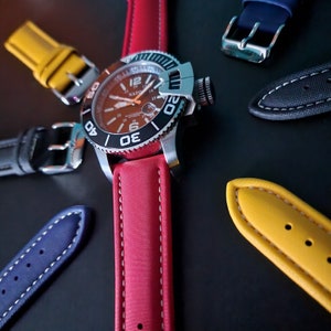 May include: A red watch strap with white stitching, a black watch face with white markings, and a silver watch case. The watch is on a black surface with other watch straps in yellow, blue, and black.