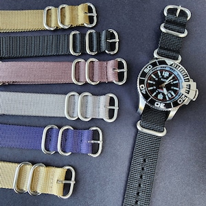 May include: Six different colored nylon watch straps with silver buckle closures. The straps are arranged vertically, with the top strap being gold, followed by black, brown, silver, navy blue, and gold. A black watch with a silver bezel and black face is shown on the right side of the image.