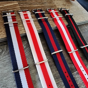 May include: Five different watch straps with red and white stripes, and one with navy blue and red stripes. The straps are made of nylon and have silver buckles.
