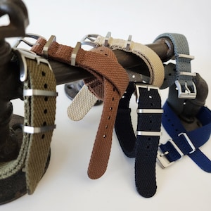 May include: A set of six watch straps in various colors, including olive green, brown, beige, navy blue, and gray. The straps are made of a woven fabric and have silver metal buckles.