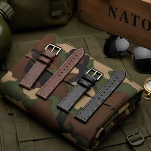 May include: Three leather watch straps in brown and black, with silver buckles and white stitching. The straps are displayed on a camouflage pattern fabric, with a wooden box labeled "NATO" in the background.